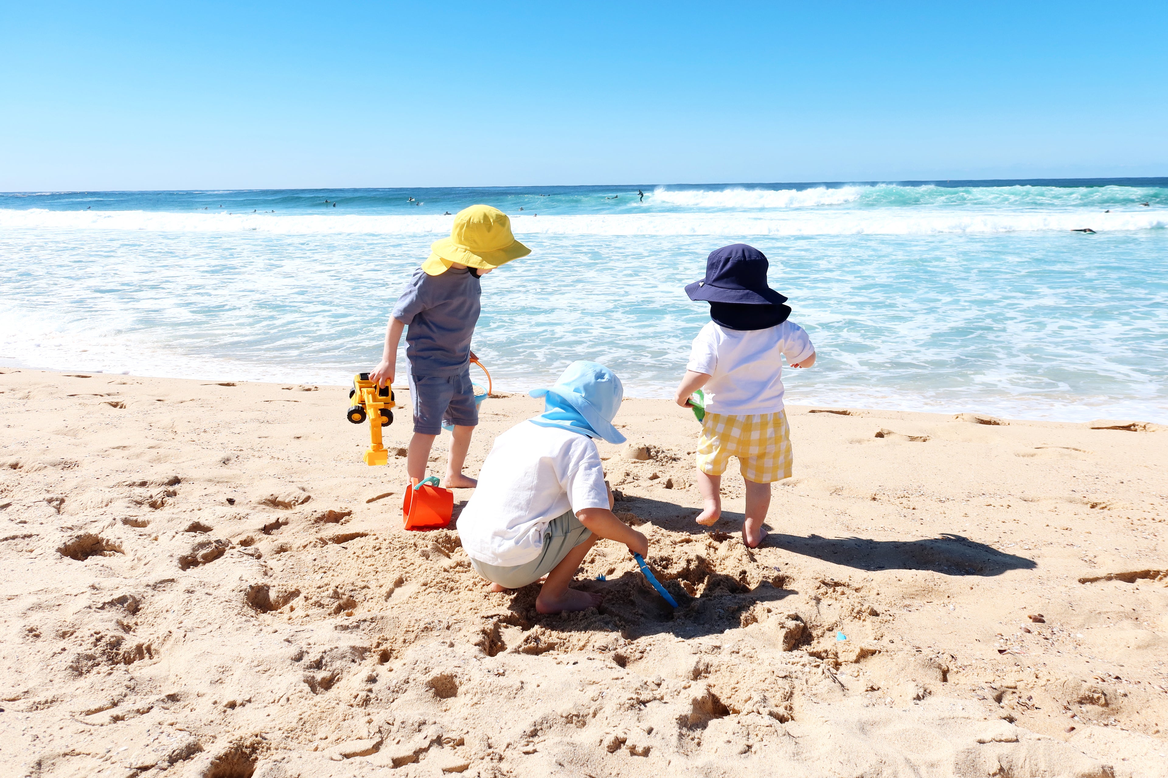 Three young children wearing Luma hats playing on a sunny beach, digging in the sand near the ocean waves